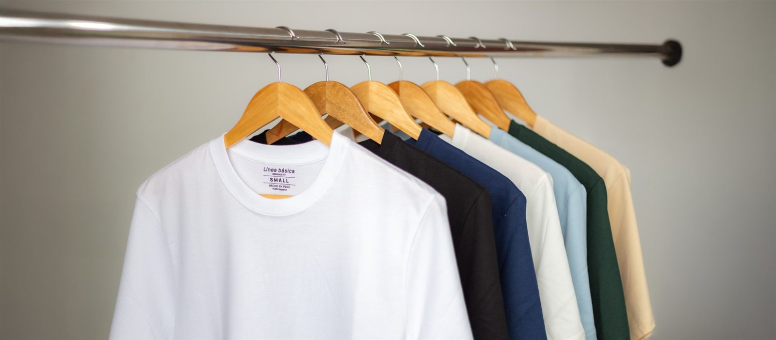 Polos Línea básica colgados en un perchero: blanco, negro, azul marino, crema, celeste, verde botella y beige